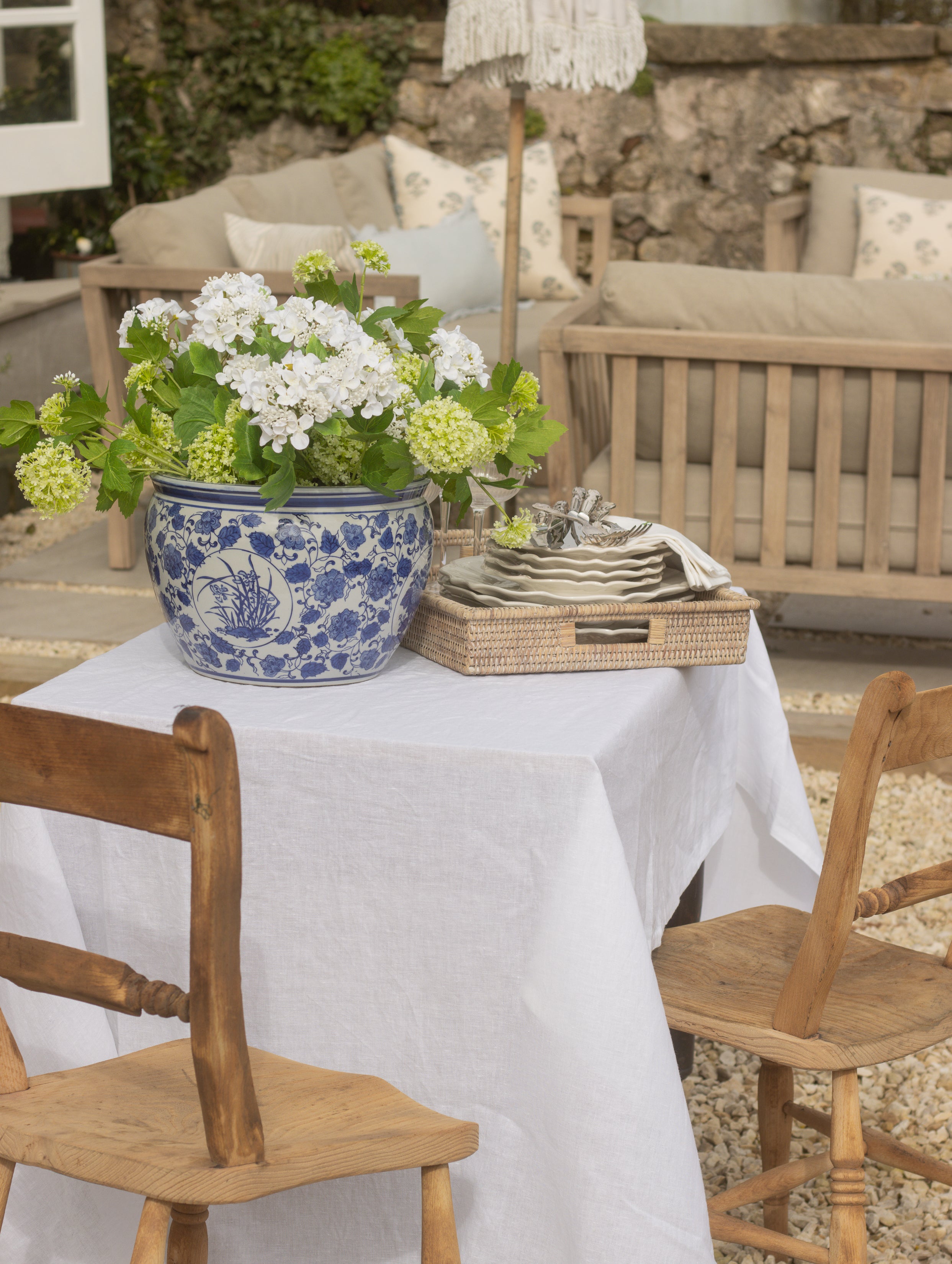 Outdoor setting with a table set for a meal, featuring a blue and white floral pot, white flowers, and wooden chairs.