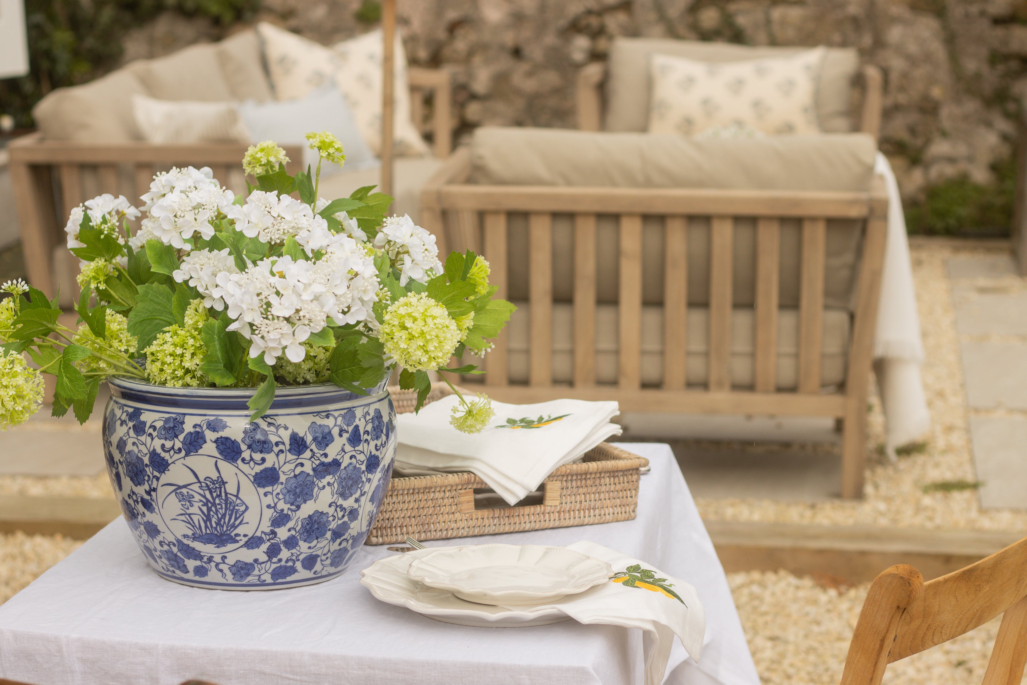 Outdoor setting with a table set for a meal, featuring a blue and white floral pot and greenery.