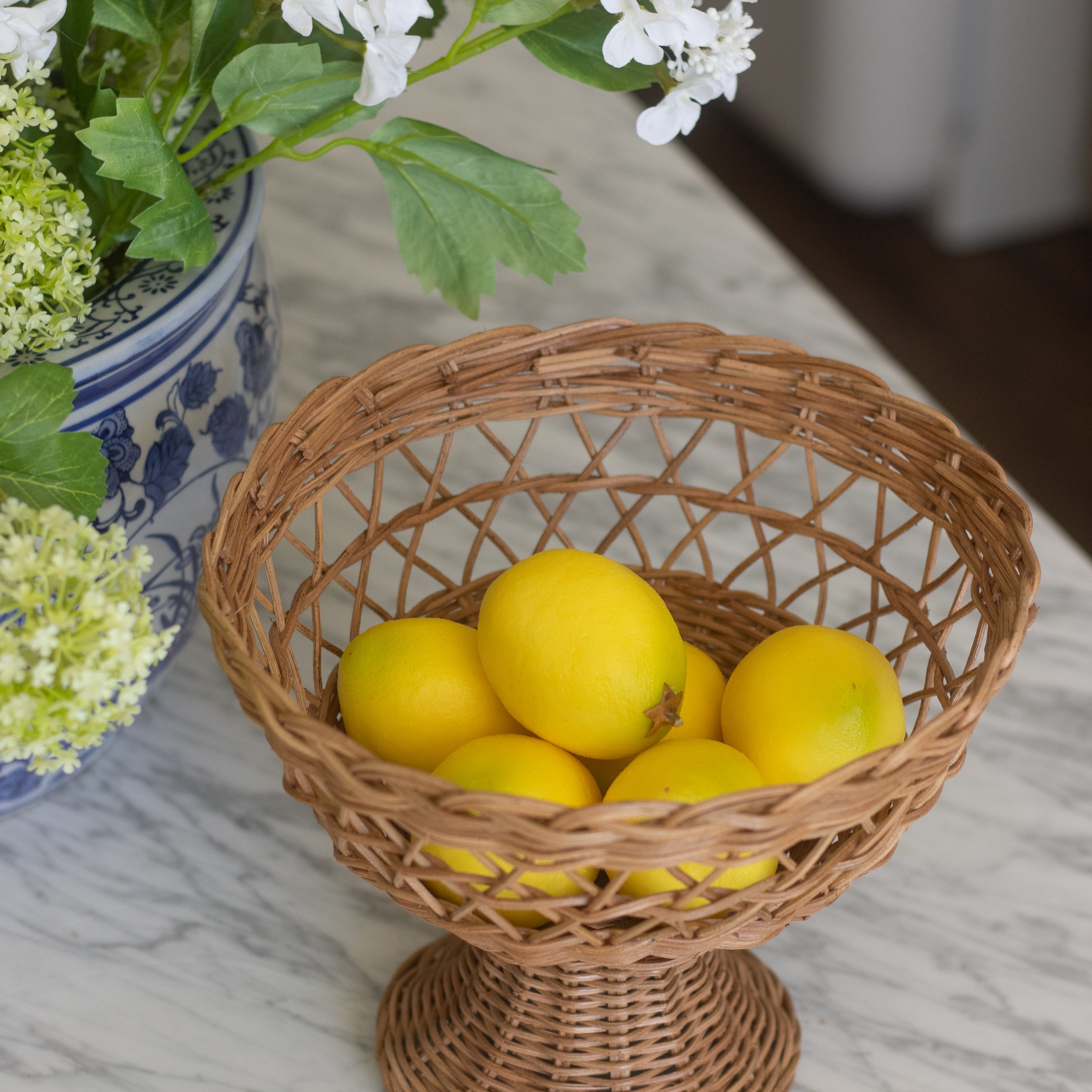 Wicker basket with lemons on a marble surface next to a vase of flowers.