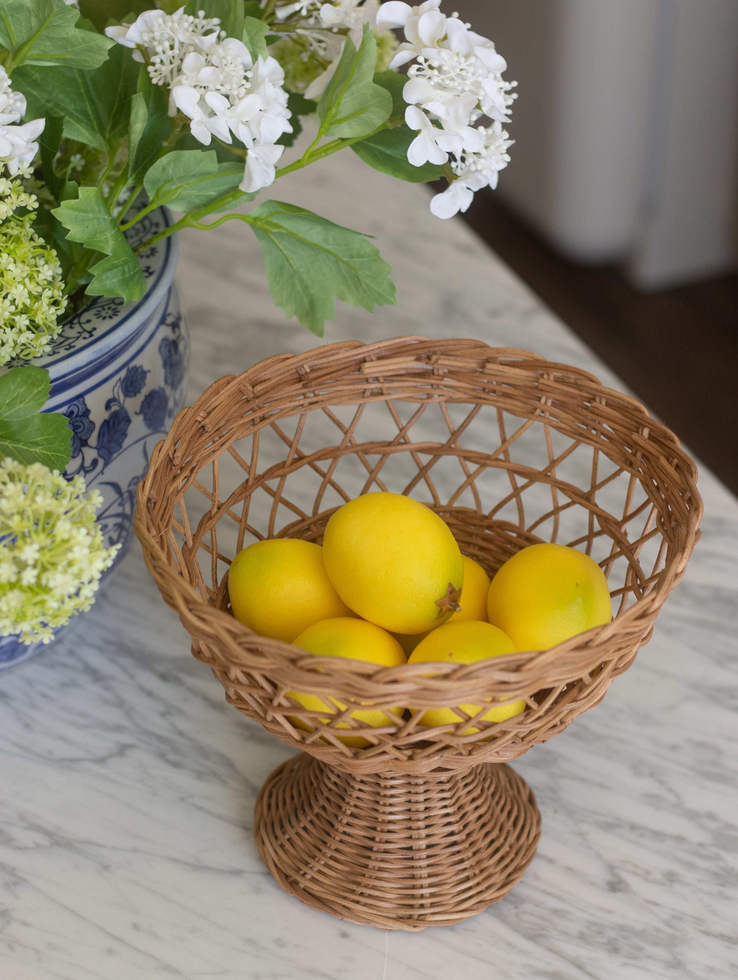 Wicker basket with lemons on a marble surface next to a vase of flowers.