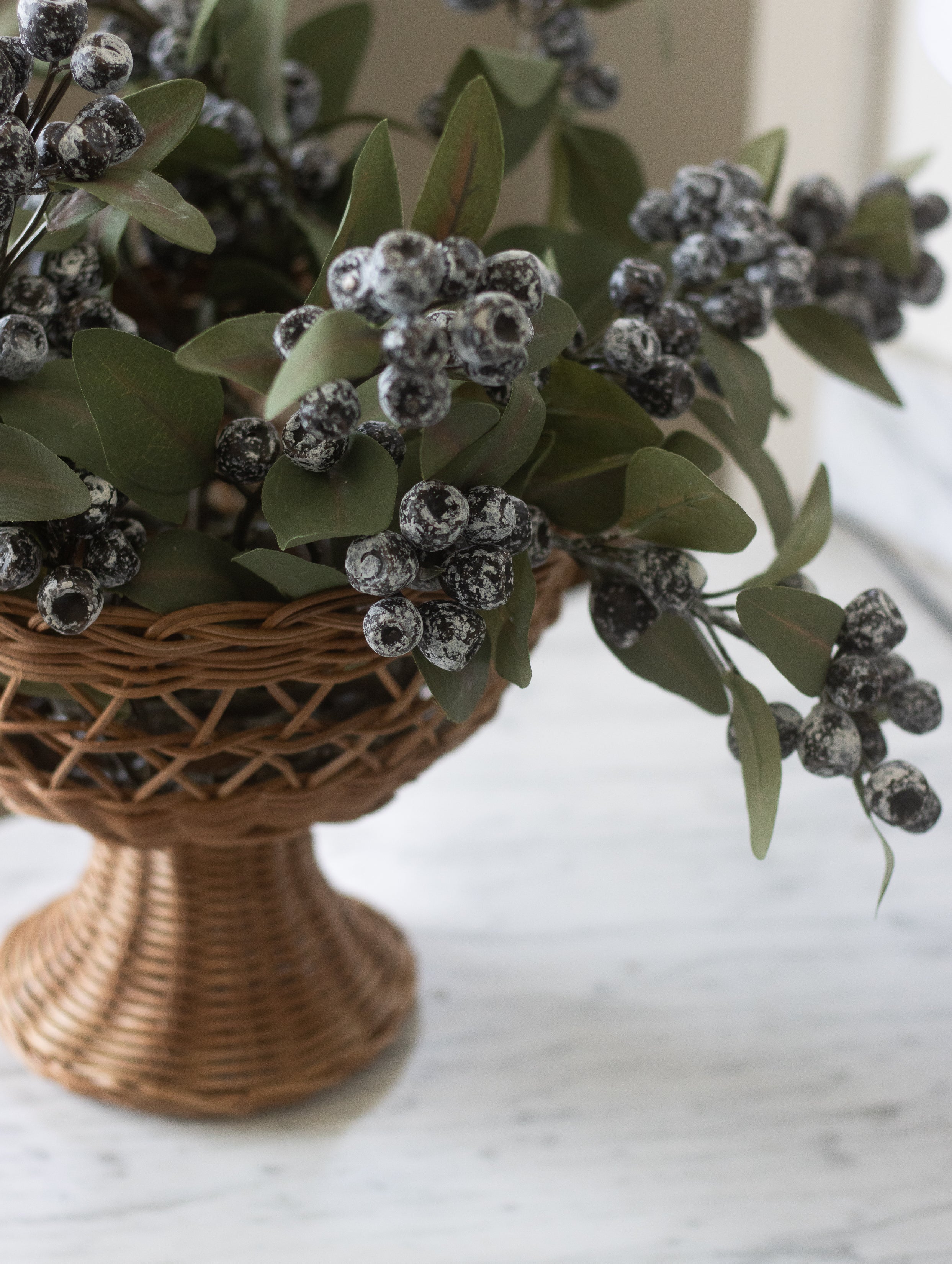 Decorative arrangement of greenery and berries in a woven basket on a marble surface.