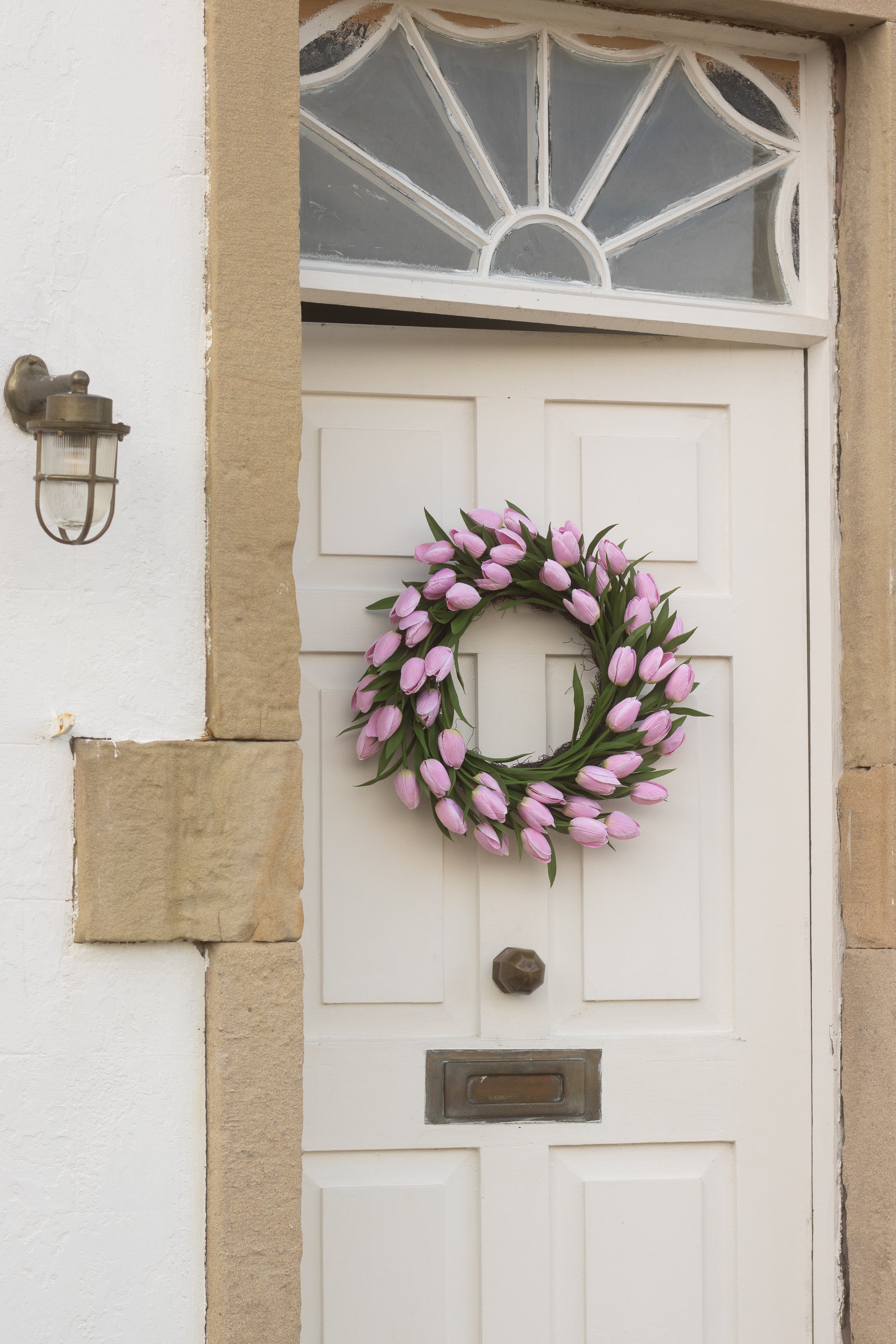 White door with a pink tulip wreath and a light fixture on a white wall.