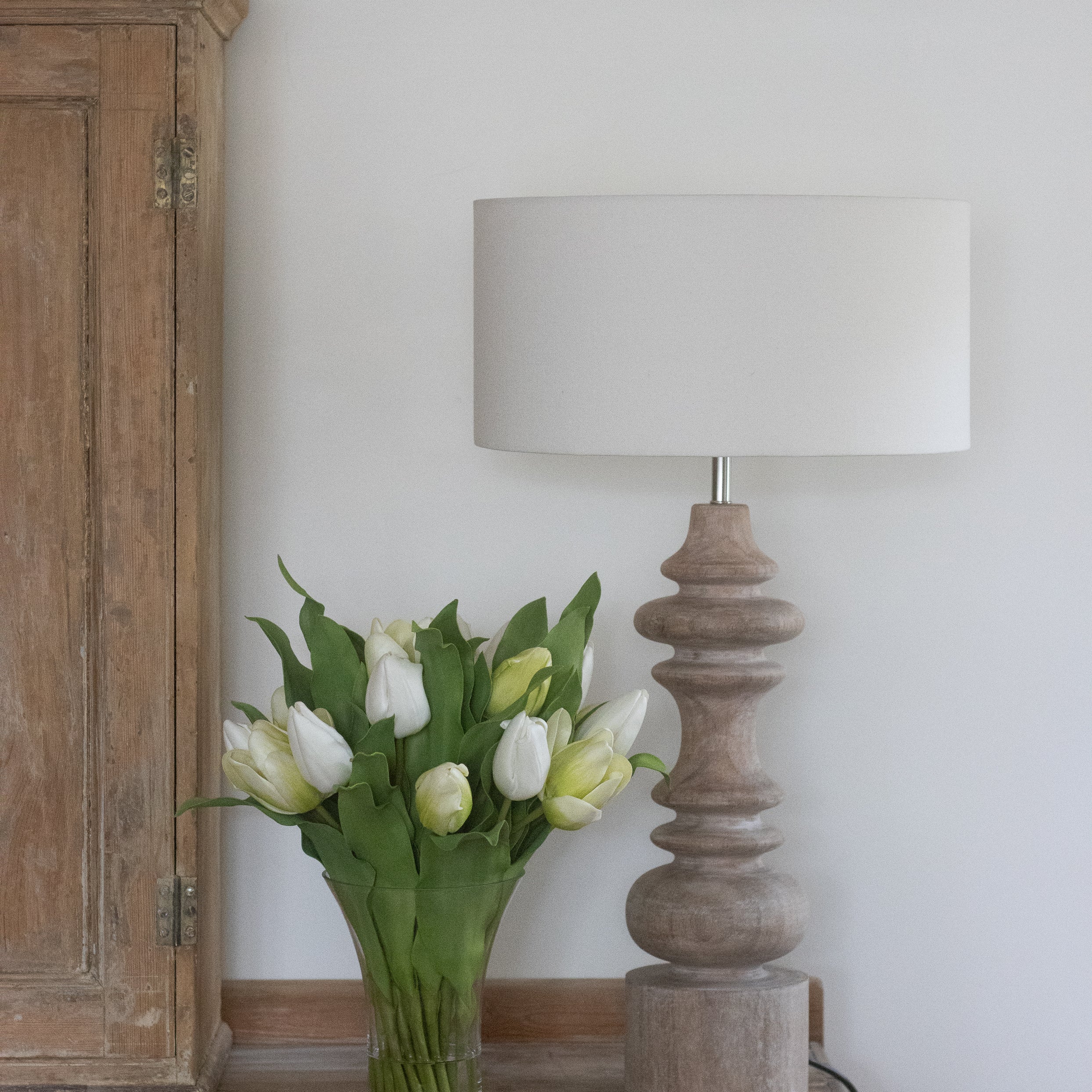 Vase of white tulips on a wooden table next to a lamp and cabinet.