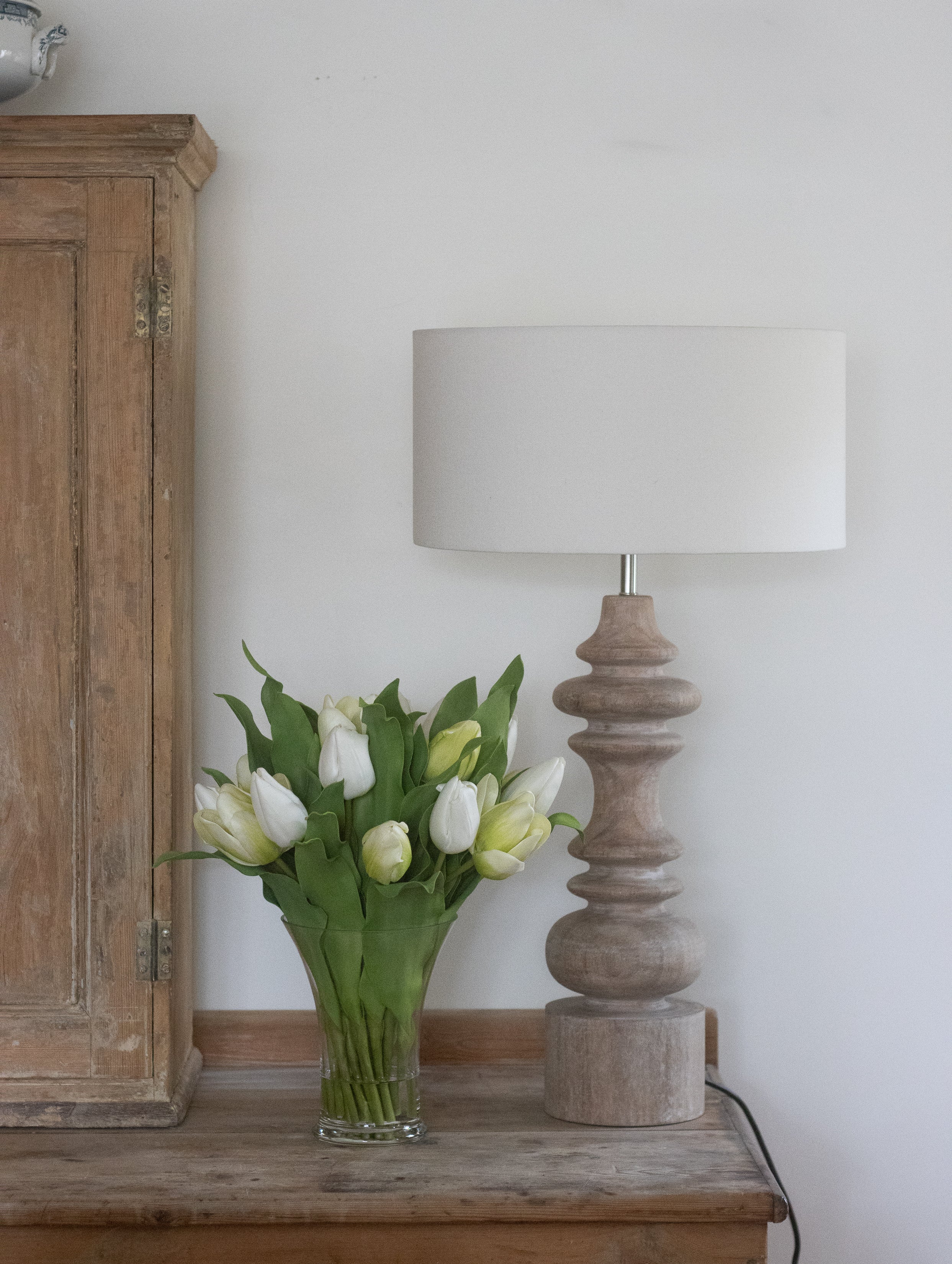 Vase of white tulips on a wooden table next to a lamp and cabinet.