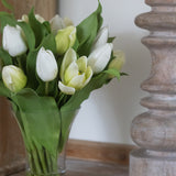 Bouquet of white and yellow tulips in a clear vase on a wooden surface with a wooden candle holder in the background.