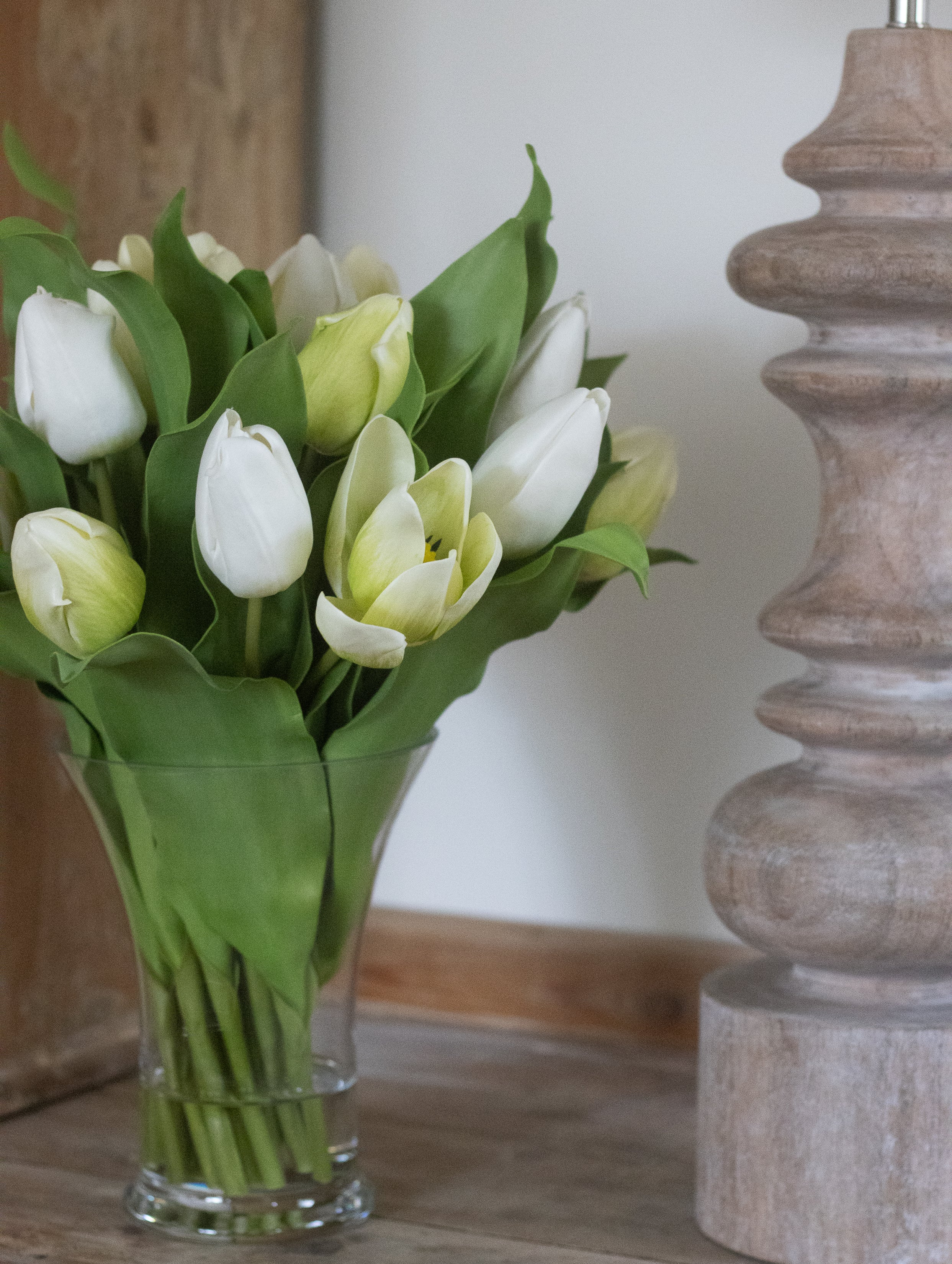 Bouquet of white and yellow tulips in a clear vase on a wooden surface with a wooden candle holder in the background.