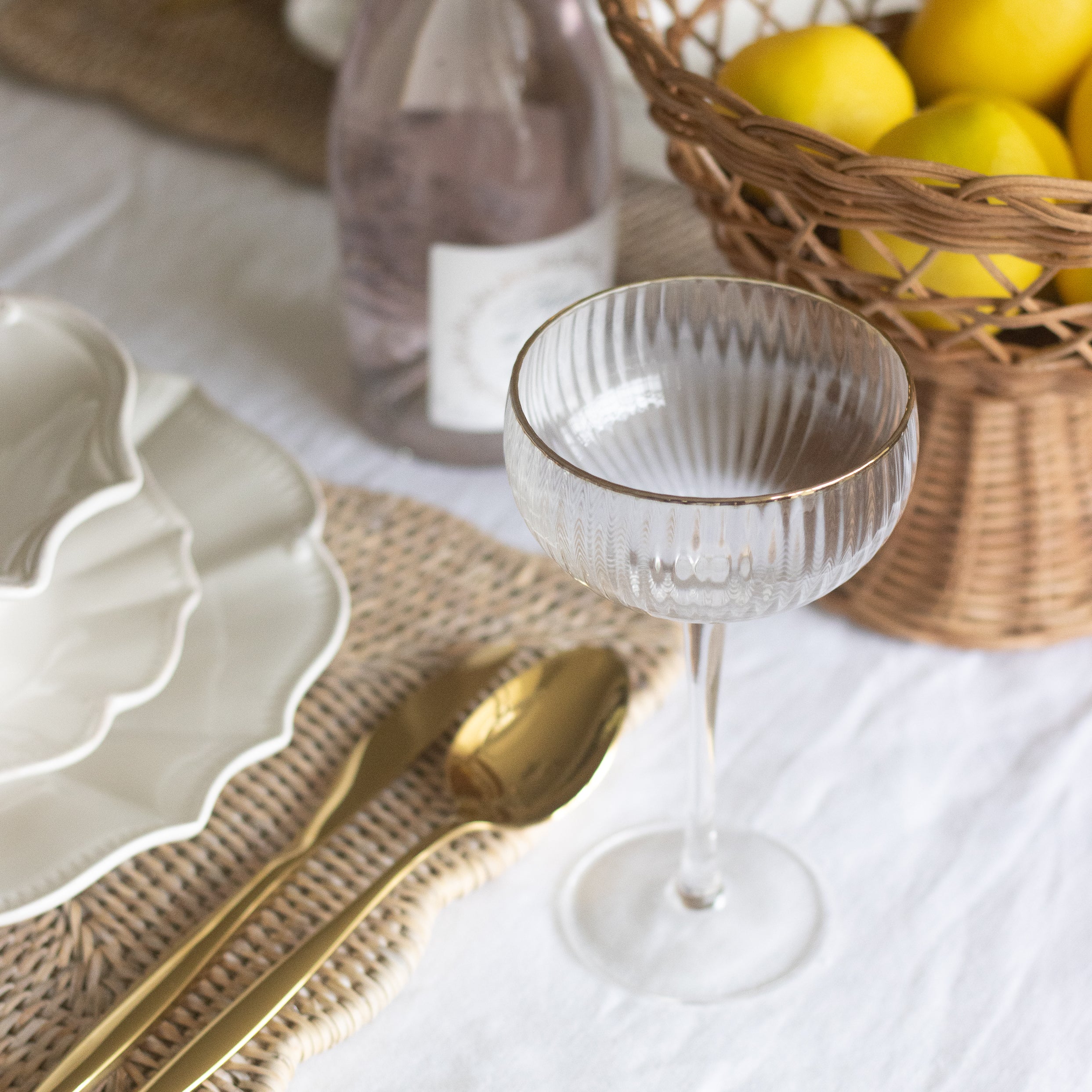Table setting with a glass, basket of lemons, and bottle of wine on a white tablecloth.