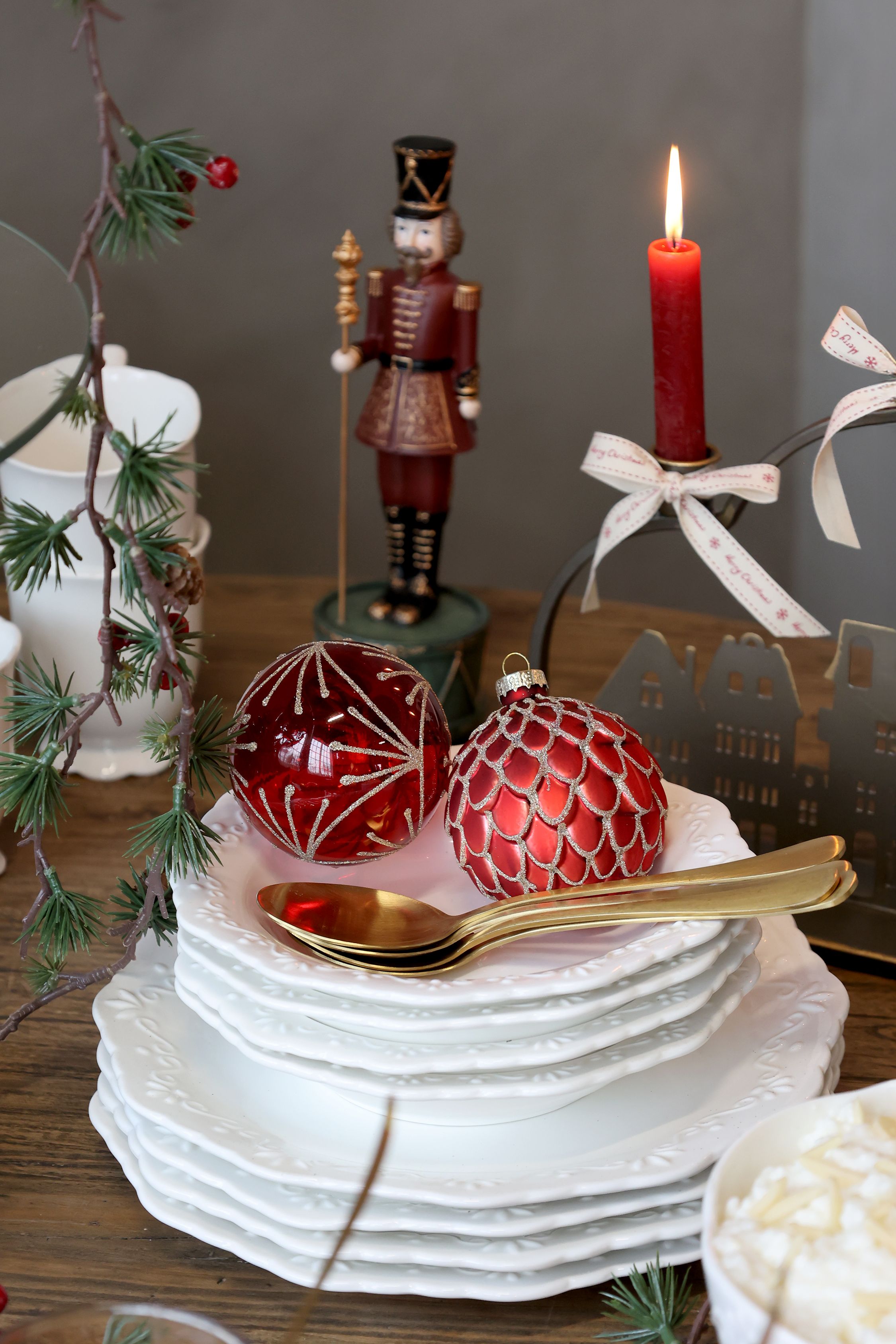 Decorative setting with red ornaments, nutcracker figurine, and lit candle on a wooden table.