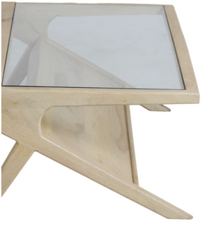 A close-up of a modern wooden table with a clear glass top and a light natural wood base featuring a sculptural, angular leg design.