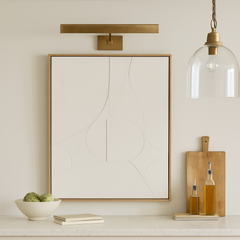 A matte brass slim picture light mounted on a wall above a white frame picture, with a pendant light hanging next to it and a wooden board with bottles on the side.