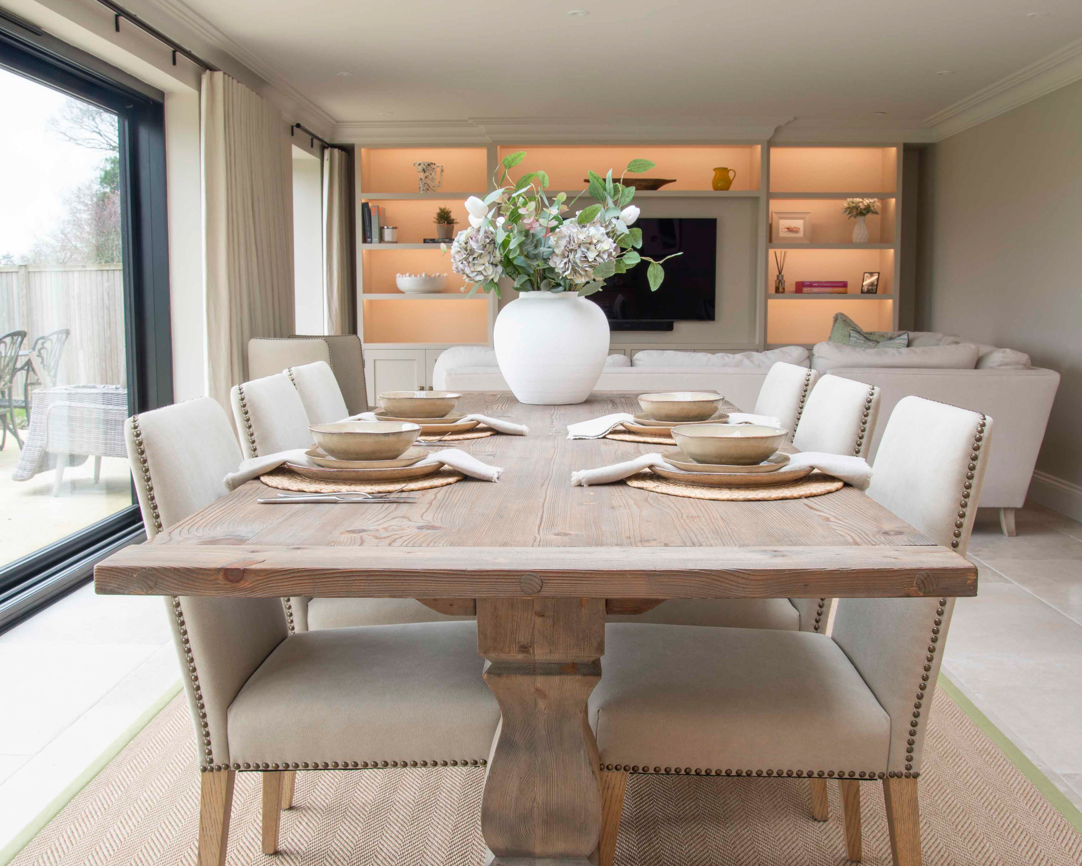 A rustic wooden dining table with a central pedestal base, surrounded by four chairs, set in a well-lit room with a large window.
