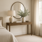 A brown oak console table with a single drawer and brass style bar handles, placed against a neutral background in a room setting with a lamp and a plant on top.