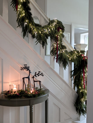 Real touch mixed evergreen garland draped up a staircase with fairy lights and red ribbon