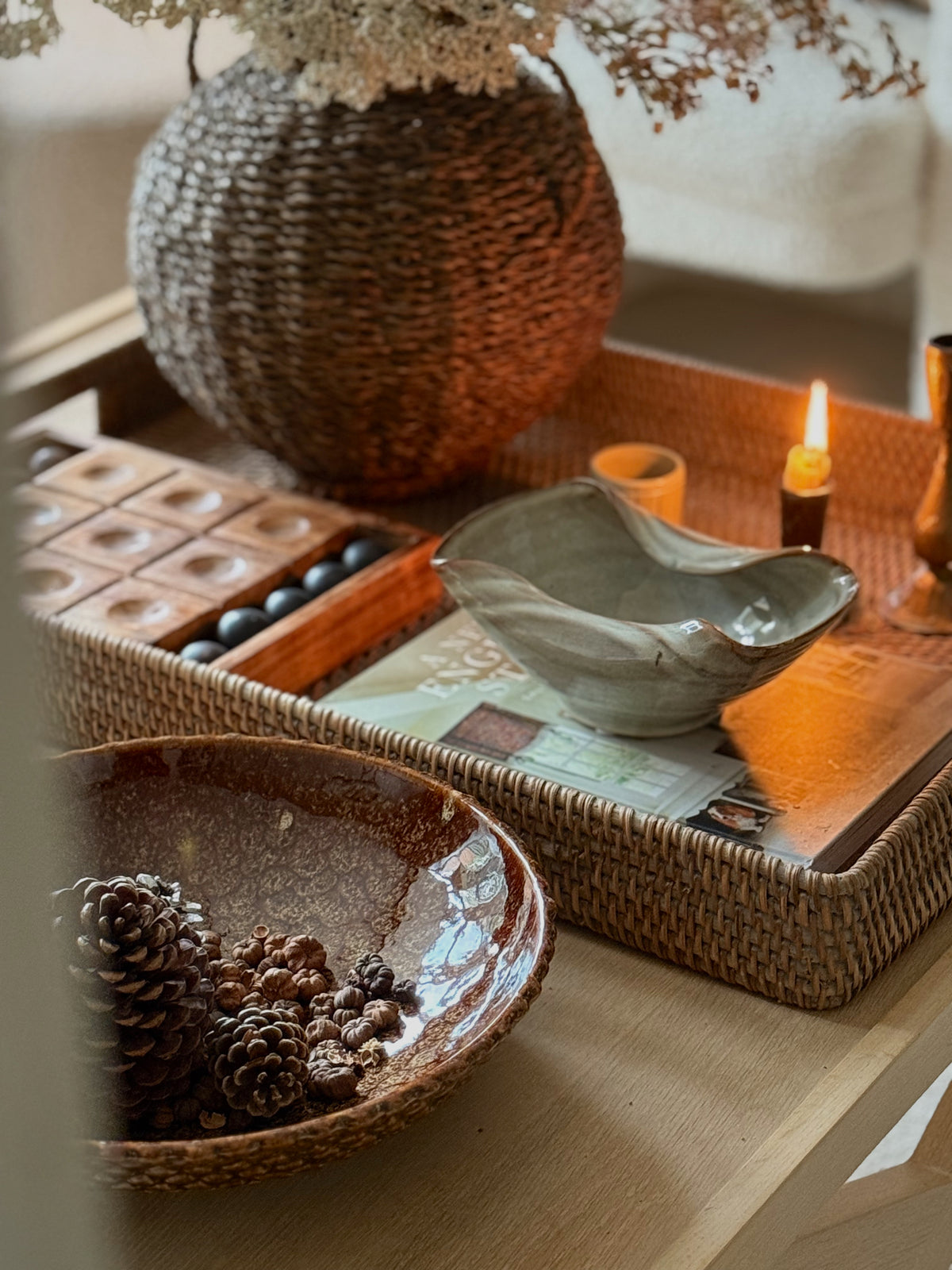 A styled tabletop scene with woven and ceramic home decor items, including a large round woven vase, a shallow ceramic bowl, a tray, candles, and a bowl of pinecones on a light wood surface.
