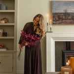Woman in a long dark dress holding flowers in a cozy living room with a fireplace.