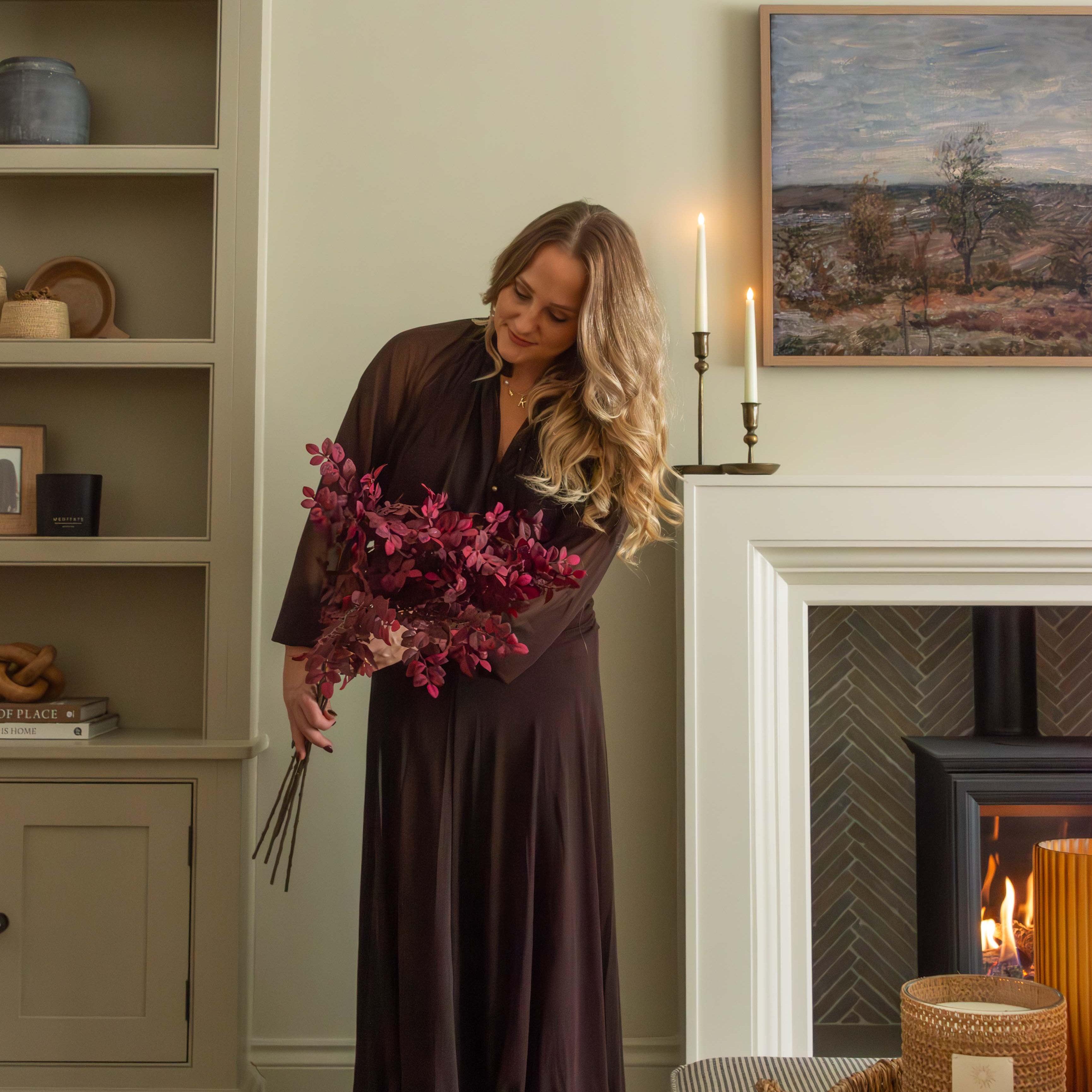 Woman in a long dark dress holding flowers in a cozy living room with a fireplace.