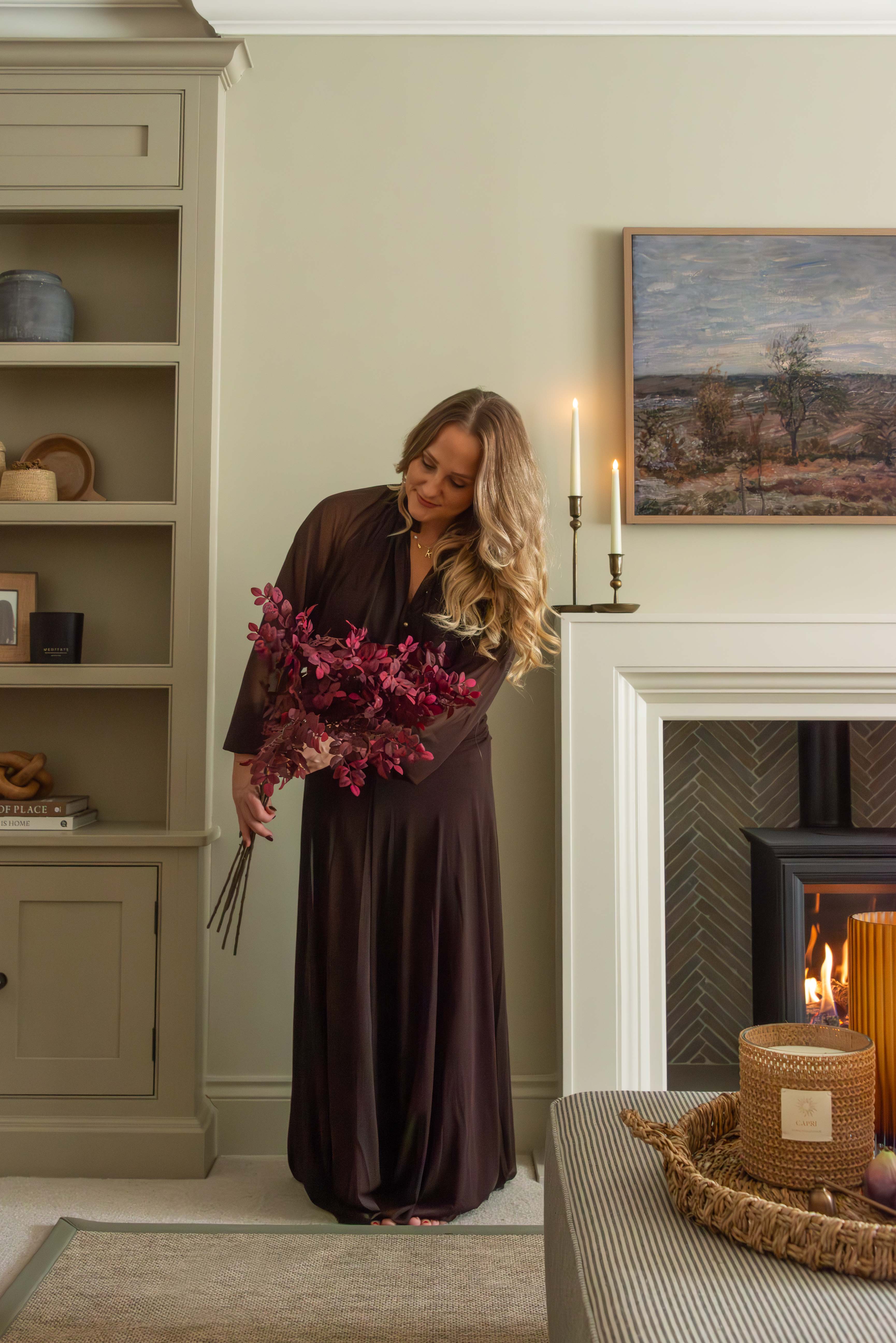 Woman in a long dark dress holding flowers in a cozy living room with a fireplace.