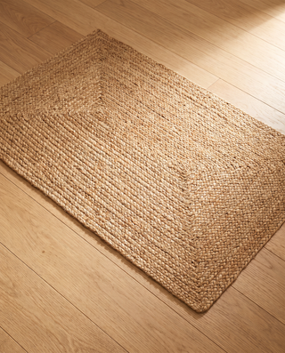 A rectangular braided jute rug in a natural tan color, shown from an elevated angle on a light-colored wooden floor.
