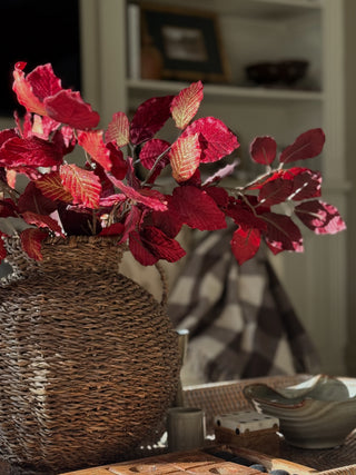 Red autumn leaves in a woven brown wicker vase, styled on a table with ceramic dishes and a plaid blanket in a cozy room setting.