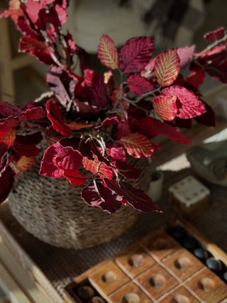 Red and burgundy faux autumn leaves in a woven basket vase, styled on a wooden table with a wooden game board in the foreground.