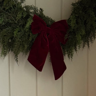 A cozy holiday-themed interior scene with a rustic wooden rocking horse on a stool, a brass trumpet decoration with a lit candle, woven storage baskets, wrapped gift boxes, a green garland with a burgundy bow, and stockings hanging on a paneled wall.