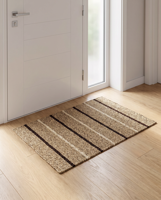 A rectangular woven entryway mat with horizontal beige and dark brown stripes, placed on a light wood floor in front of a white door.