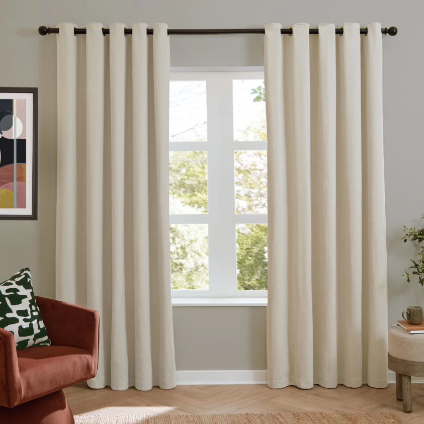 Beige curtains hanging in a room with a window, chair, and abstract art on the wall.