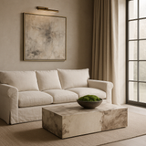A marble coffee table with a neutral tone, placed in a living room setting with a beige sofa, a framed abstract picture on the wall, and large windows.