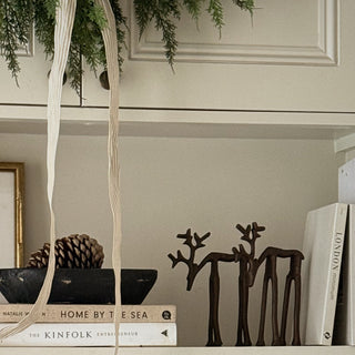 A styled interior shelf scene with a dark metal bowl, pinecone, books, and two dark wooden deer-shaped bookends holding a white book on a cream shelf.