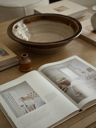 A rustic brown ceramic bowl sits on a light wood table next to an open magazine and a small brown vase, in a softly styled interior setting.