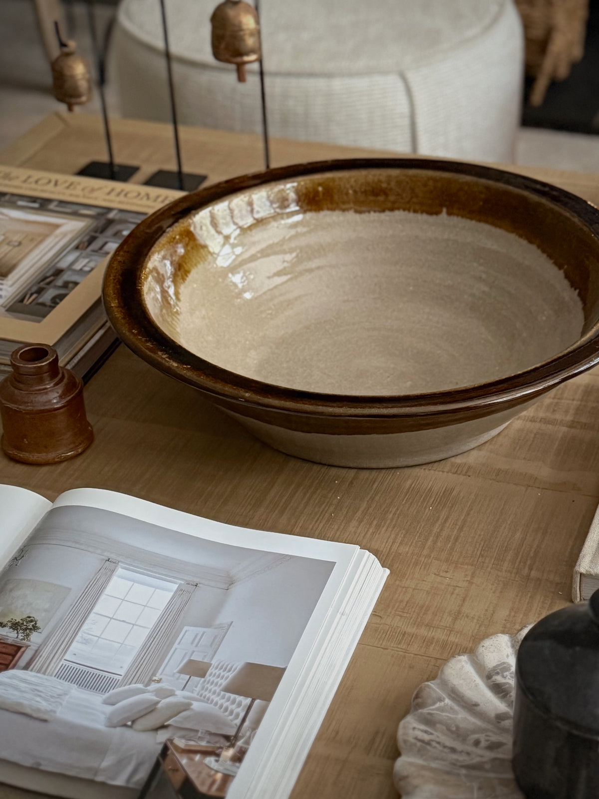 A large round ceramic bowl with a rustic brown rim and a warm beige glazed interior, styled on a wooden table with an open book, small amber bottle, and other home decor items.
