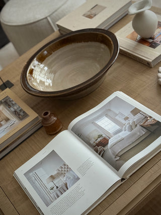 A styled interior scene showing a large round ceramic bowl on a wooden table, alongside an open magazine with bedroom photography, stacked books, a small amber bottle, and a white vase.