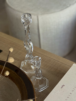 Two clear twisted glass candlestick holders on a light wood table, with a neutral fabric stool in the background and a decorative plate partially visible in the foreground.