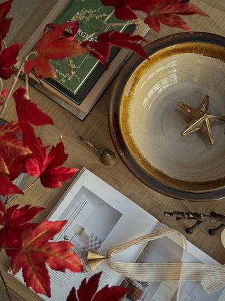 A styled flat-lay featuring a large ceramic bowl with a gold star ornament inside, surrounded by red maple leaves, a book, and an open magazine on a wooden surface.