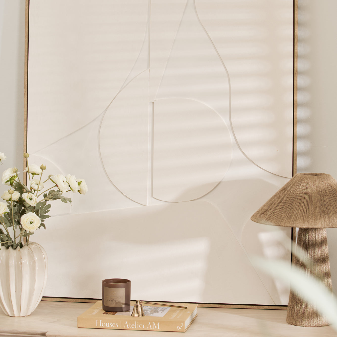 An abstract painting in a wooden frame displayed on a wooden dresser, with a vase of flowers and a lamp beside it.