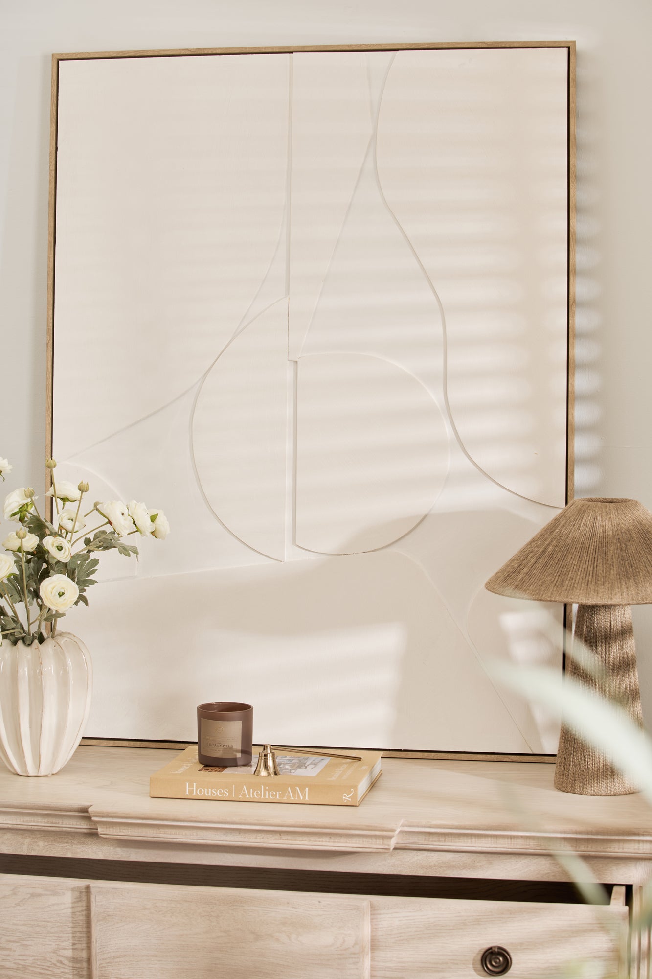 An abstract painting in a wooden frame displayed on a wooden dresser, with a vase of flowers and a lamp beside it.