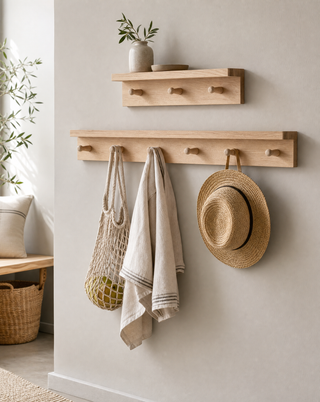A light natural wood wall-mounted coat rack with two hooks and a smaller matching shelf above, styled in a bright interior with towels, a straw hat, and a woven bag hanging from the hooks.