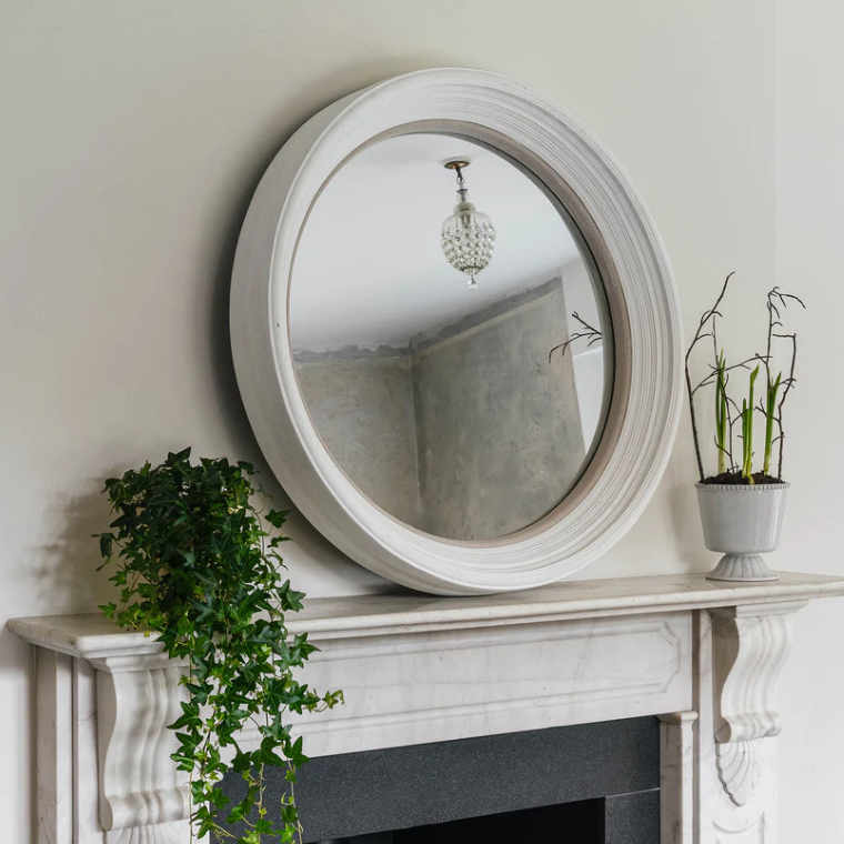 Round mirror with white frame on a fireplace mantel with plants.