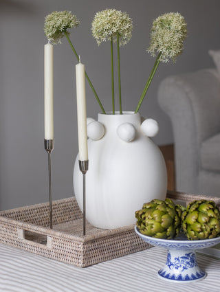 White vase with flowers and candles on a tray, with artichokes on a small stand in the background.