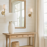A light oak console table with turned legs stands against a white shiplap wall in a bright living room. Above it hangs a matching wood-framed mirror, flanked by two brass wall sconces with white shades. On the table sits a large cream ceramic lamp with a white shade, while a woven basket rests on the lower shelf. To the right, long beige linen curtains frame a large window letting in natural light, and light wood flooring completes the airy, neutral-toned space.