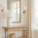 A light oak console table with turned legs stands against a white shiplap wall in a bright living room. Above it hangs a matching wood-framed mirror, flanked by two brass wall sconces with white shades. On the table sits a large cream ceramic lamp with a white shade, while a woven basket rests on the lower shelf. To the right, long beige linen curtains frame a large window letting in natural light, and light wood flooring completes the airy, neutral-toned space.