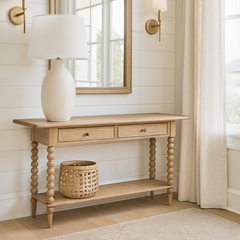 A light oak console table with turned legs stands against a white shiplap wall in a bright living room. Above it hangs a matching wood-framed mirror, flanked by two brass wall sconces with white shades. On the table sits a large cream ceramic lamp with a white shade, while a woven basket rests on the lower shelf. To the right, long beige linen curtains frame a large window letting in natural light, and light wood flooring completes the airy, neutral-toned space.