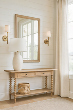 A light oak console table with turned legs stands against a white shiplap wall in a bright living room. Above it hangs a matching wood-framed mirror, flanked by two brass wall sconces with white shades. On the table sits a large cream ceramic lamp with a white shade, while a woven basket rests on the lower shelf. To the right, long beige linen curtains frame a large window letting in natural light, and light wood flooring completes the airy, neutral-toned space.