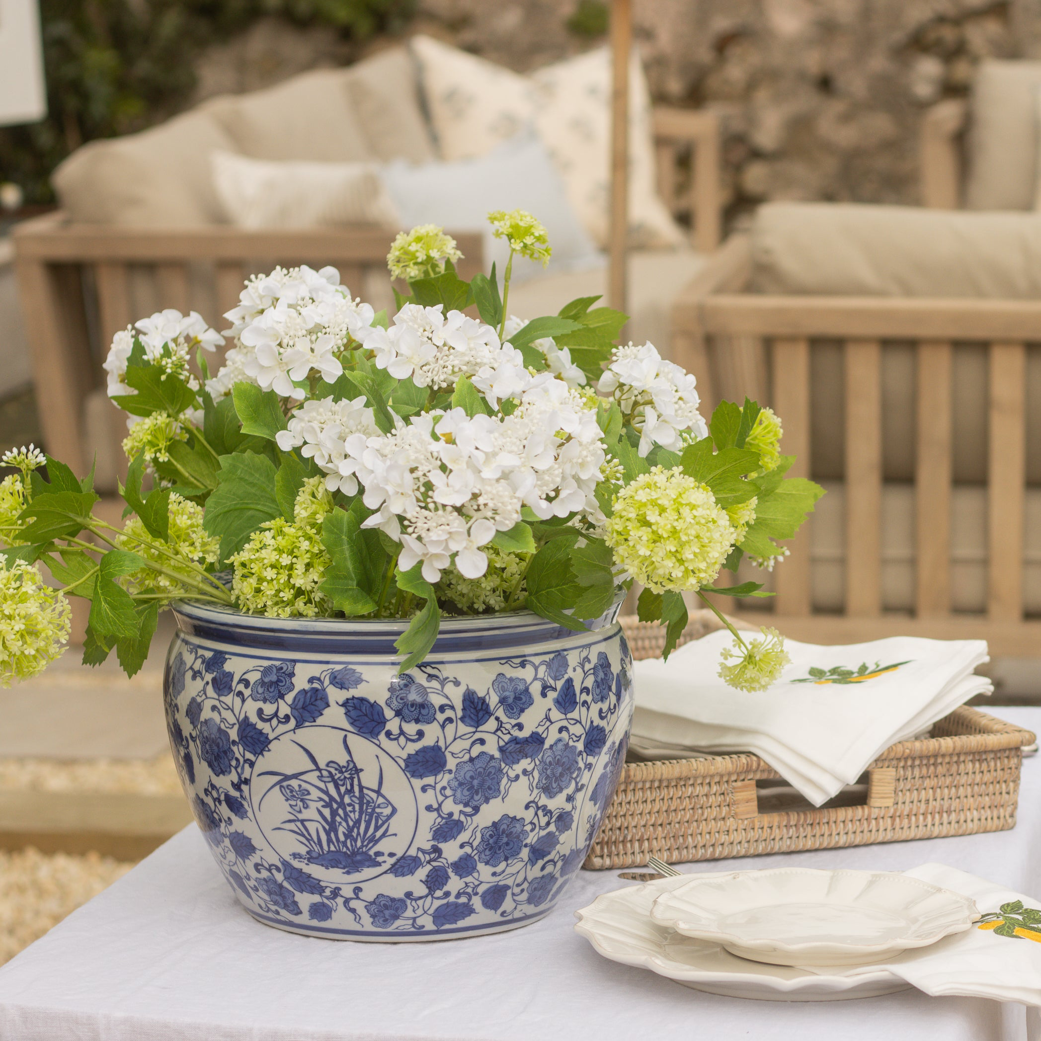 Outdoor setting with a table set for a meal, featuring a blue and white floral pot and greenery.