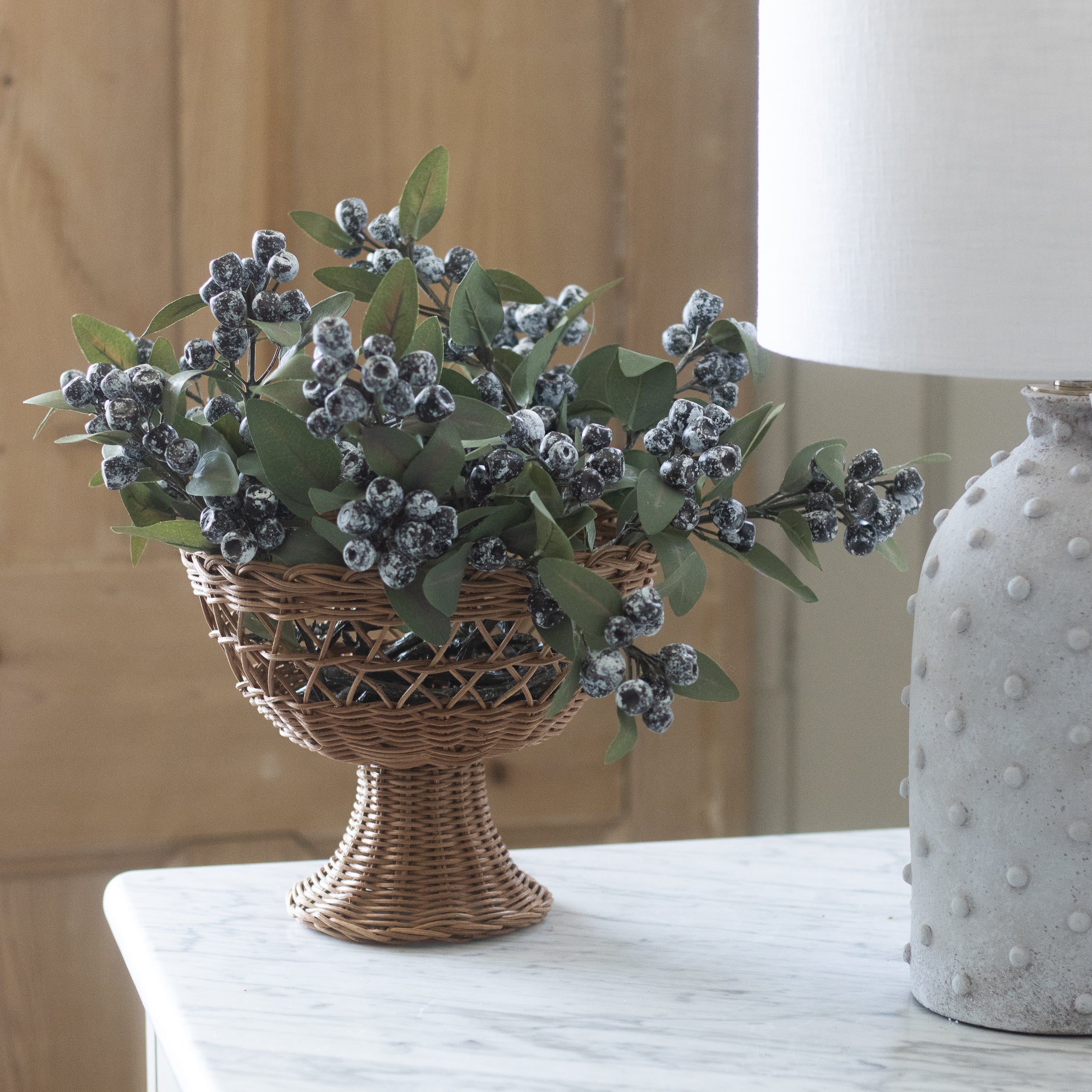 Decorative faux berries in a woven basket on a wooden surface with a lamp in the background.