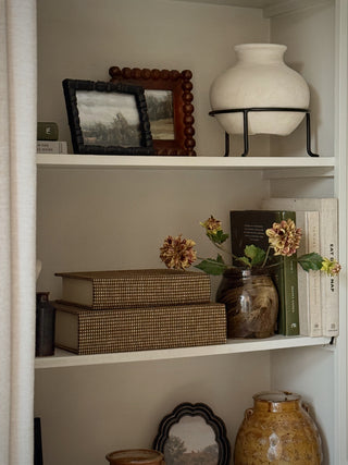 A styled interior shelf scene with white shelving holding a large white ceramic vase on a black metal stand, framed art, books, stacked decorative boxes, and a small brown vase with dried flowers.