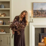 Woman in a long dark dress holding flowers in a cozy room with candles and a fireplace.