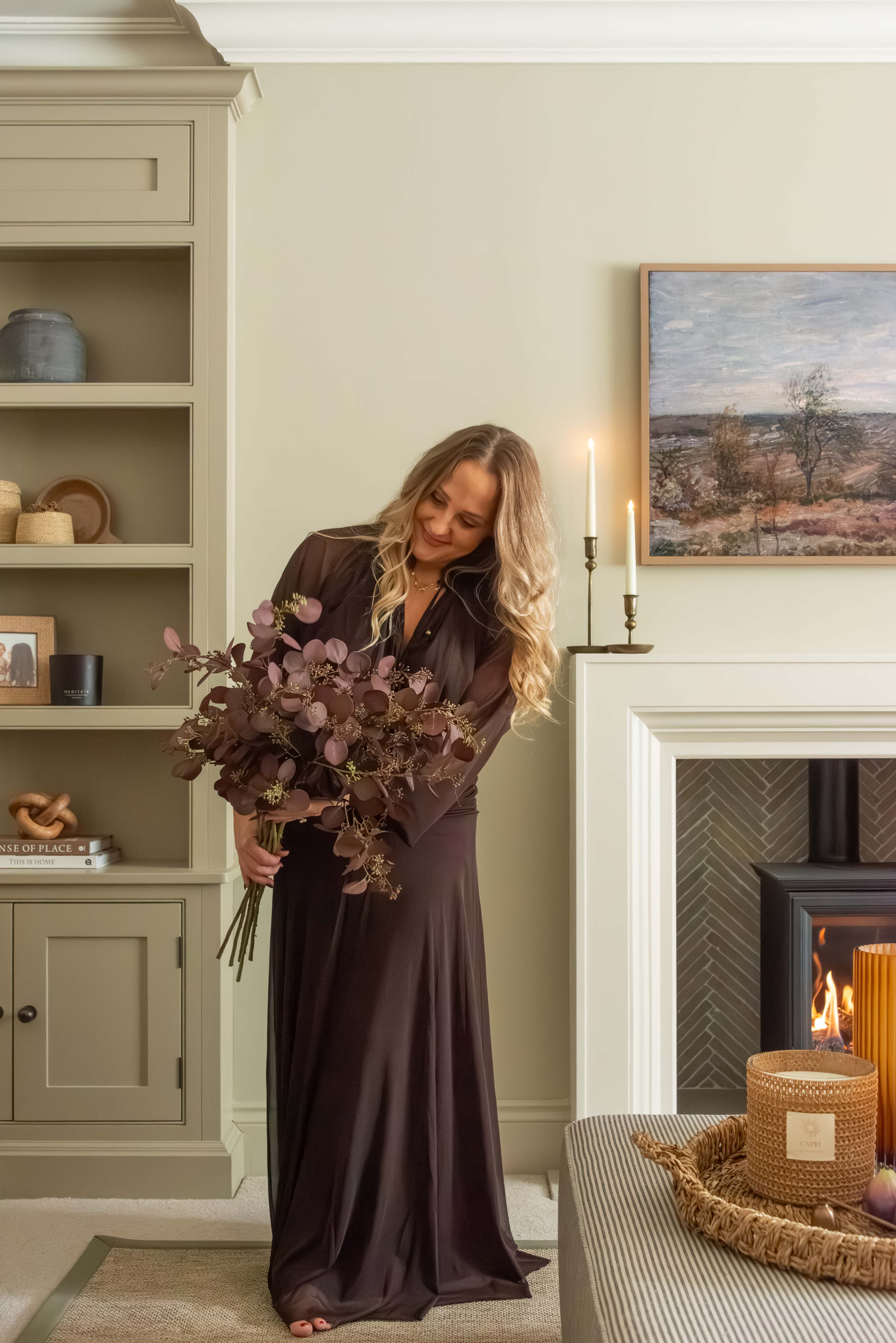 Woman in a long dark dress holding flowers in a cozy room with candles and a fireplace.