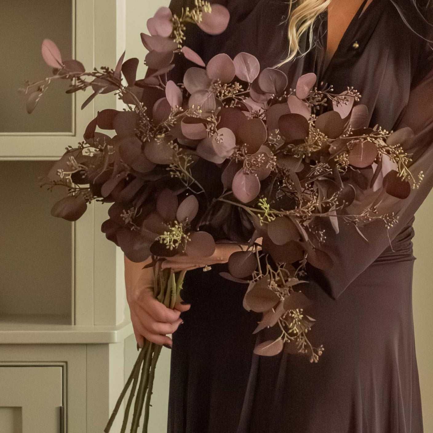 Woman in a long dark dress holding flowers in a cozy room with candles and a fireplace.
