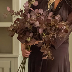 Woman in a long dark dress holding flowers in a cozy room with candles and a fireplace.